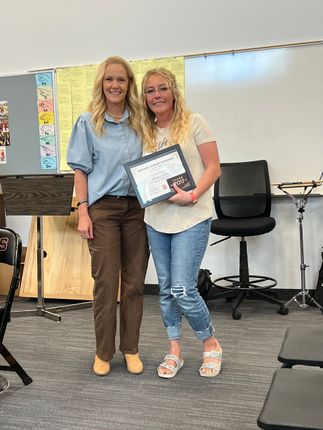 Two women stand in a classroom, smiling and holding a framed certificate.