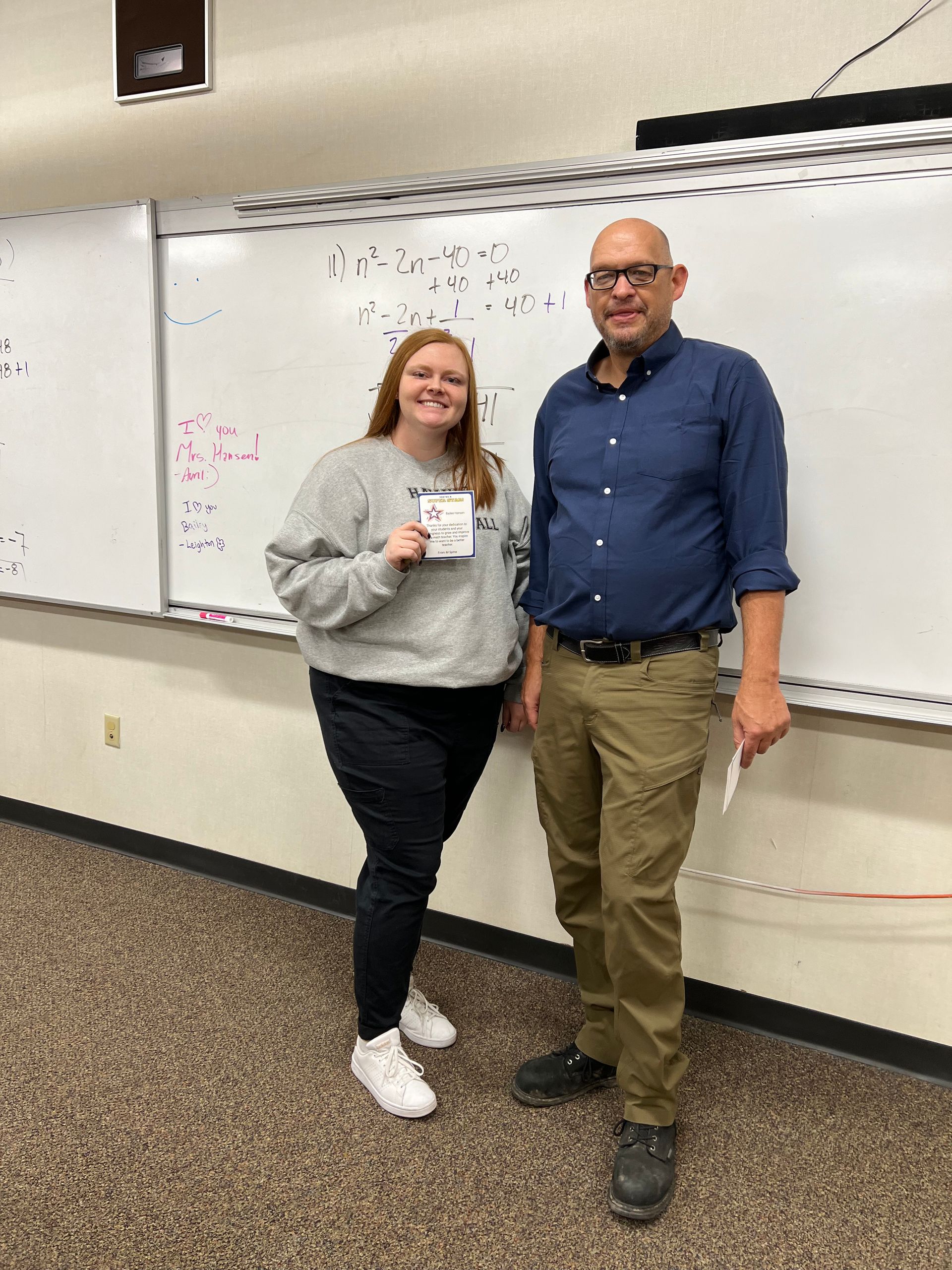 Woman in a grey sweatshirt and man in a blue shirt stand next to a whiteboard. The woman is holding a card.