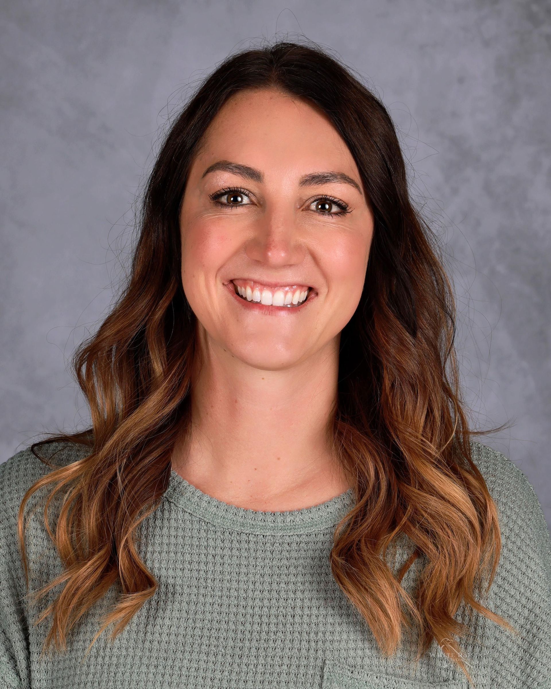 A smiling person with long, wavy brown hair wearing a light green textured sweater against a mottled gray background.