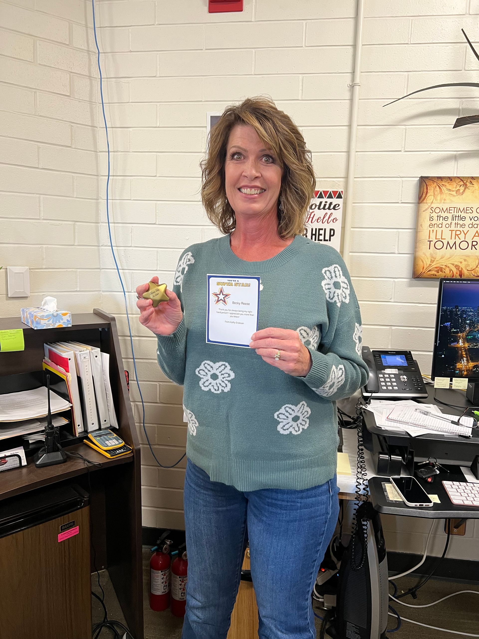 Woman holding certificate and small award in office; wearing a green sweater and jeans.