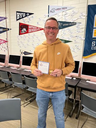 A person in a yellow hoodie and jeans stands in a classroom, holding a small card and smiling at the camera.