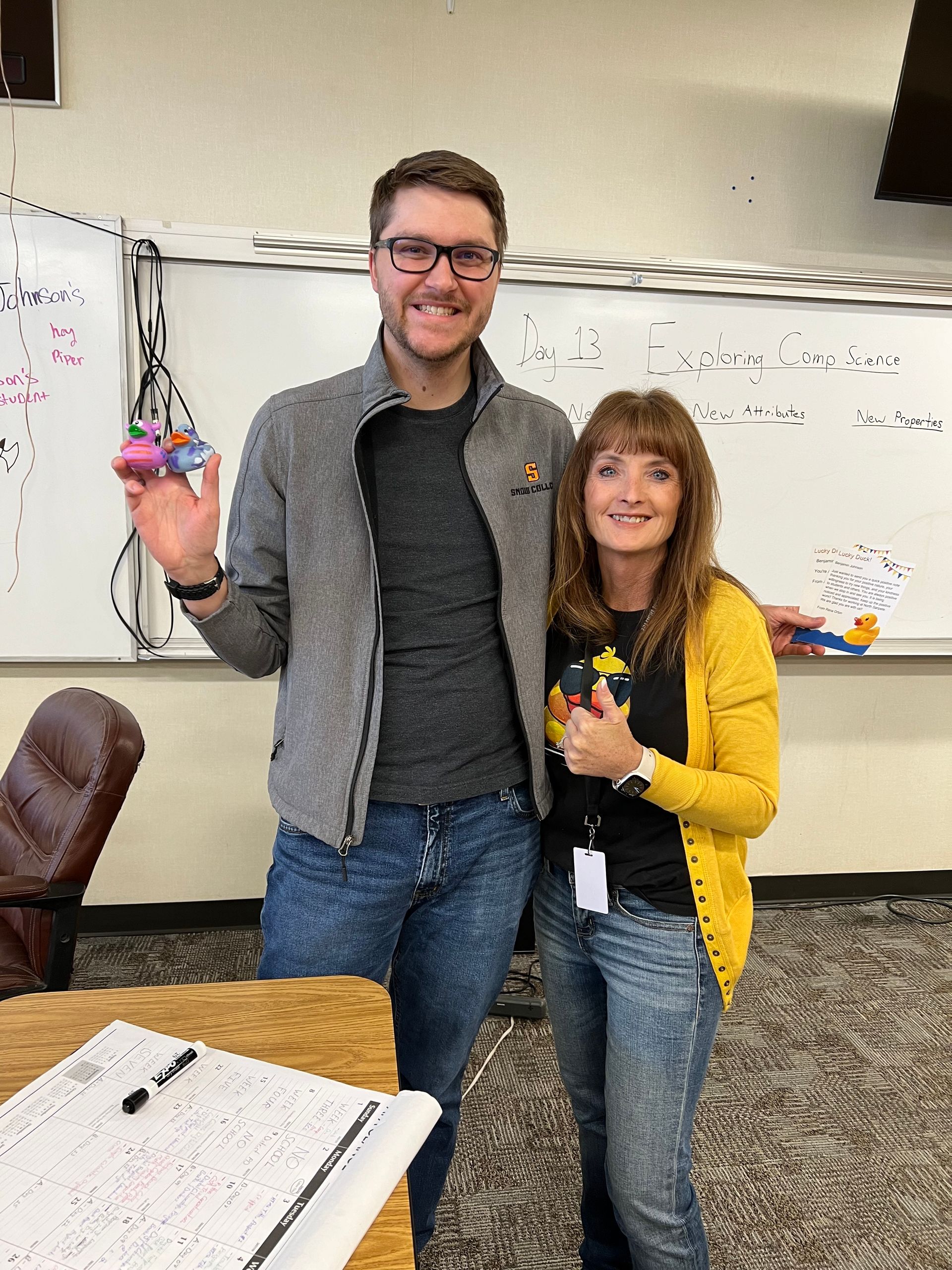 A man and a woman are posing for a picture in front of a whiteboard in a classroom.