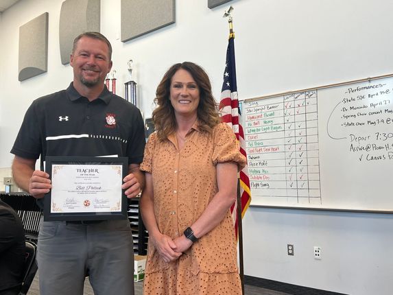 Man holding a certificate beside a woman in an office, with an American flag and whiteboard behind them.
