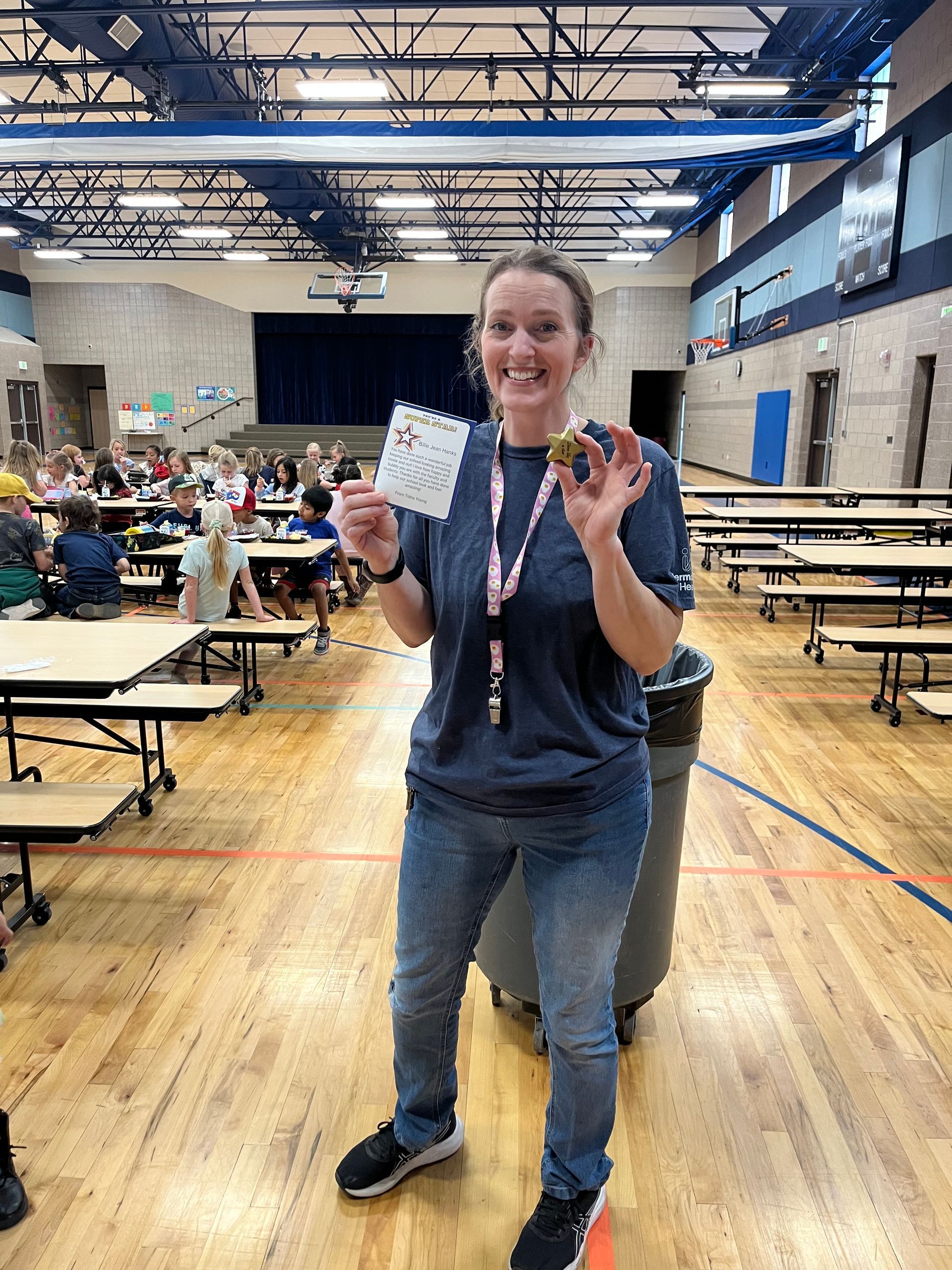 Woman in gymnasium holding a lanyard and small cards, smiling on a wooden court with tables and people behind her.