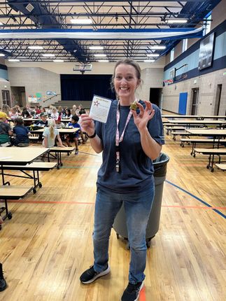 Person smiling and holding a card and medal in a gymnasium with tables and people in the background