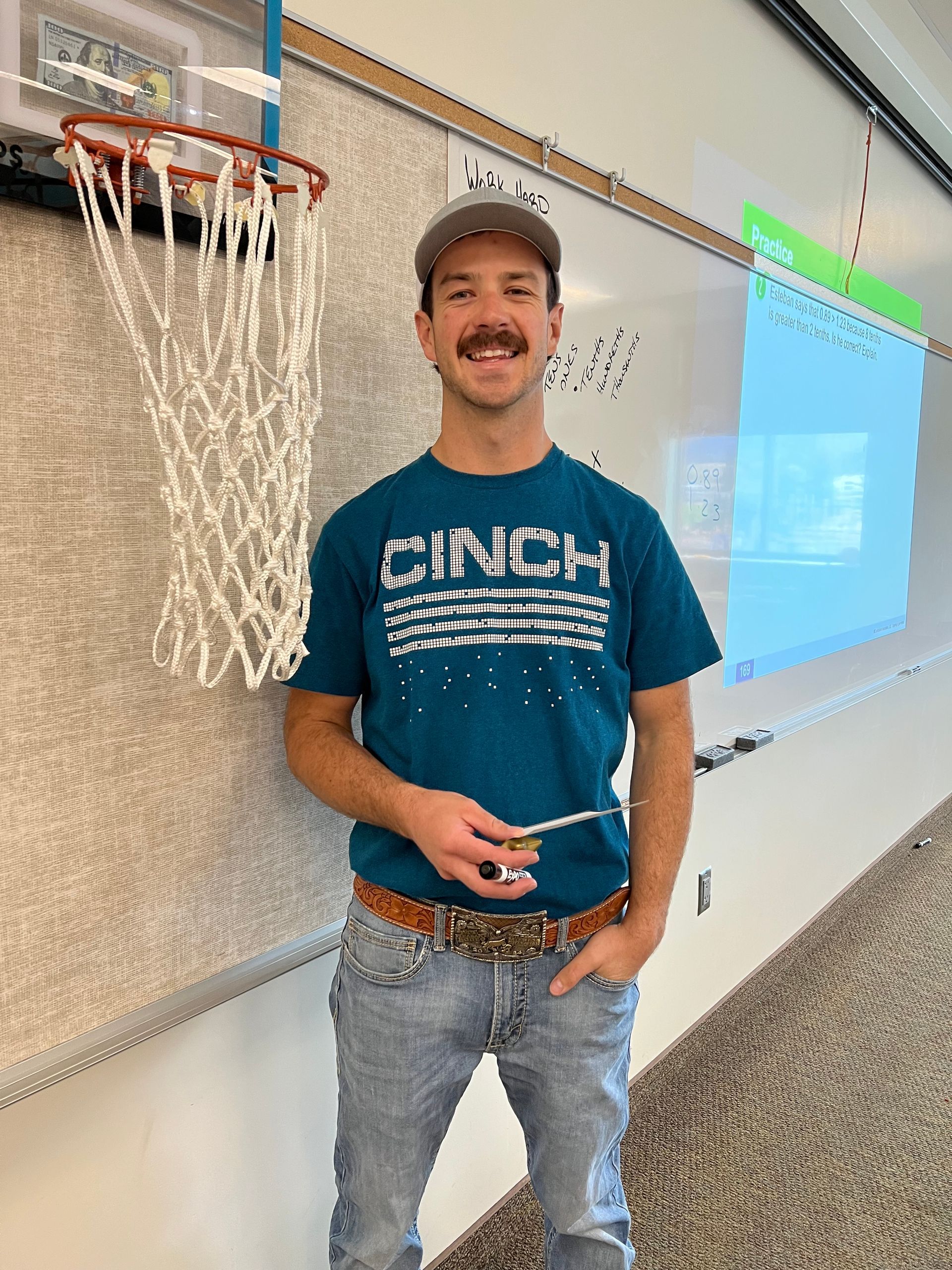 Man wearing a teal shirt, jeans, and a hat stands in front of a whiteboard and basketball hoop.