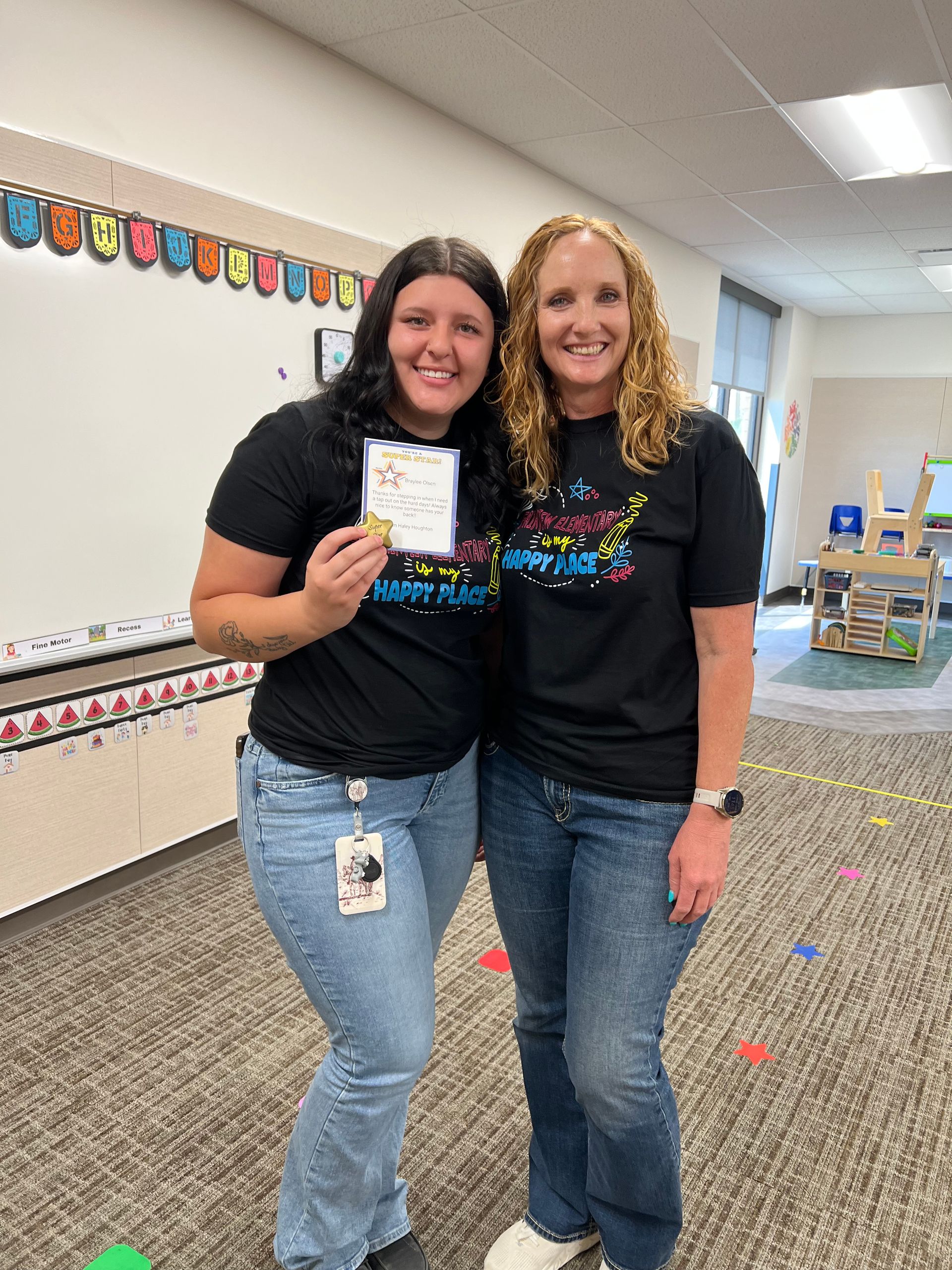 Two smiling coworkers in black shirts stand together in a classroom, one holding a small card.