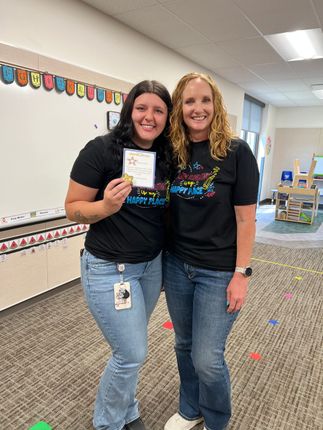 Two smiling teachers in black shirts pose in a classroom, one holding a small award.