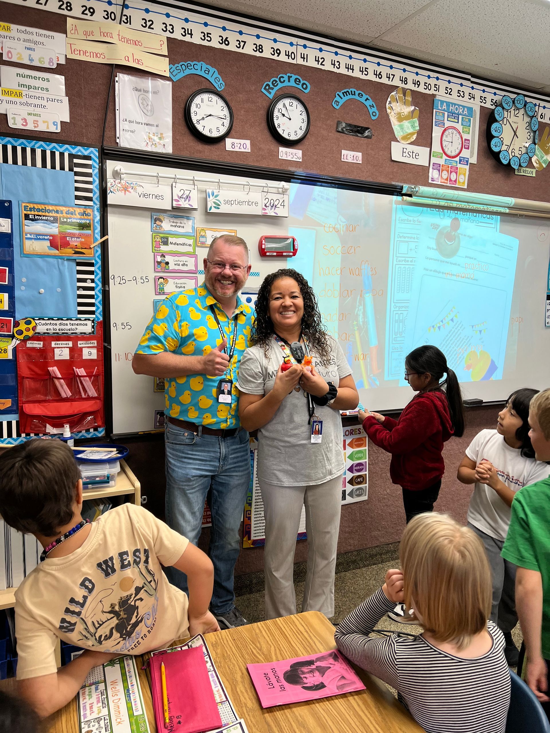 A man and a woman are standing in front of a group of children in a classroom.