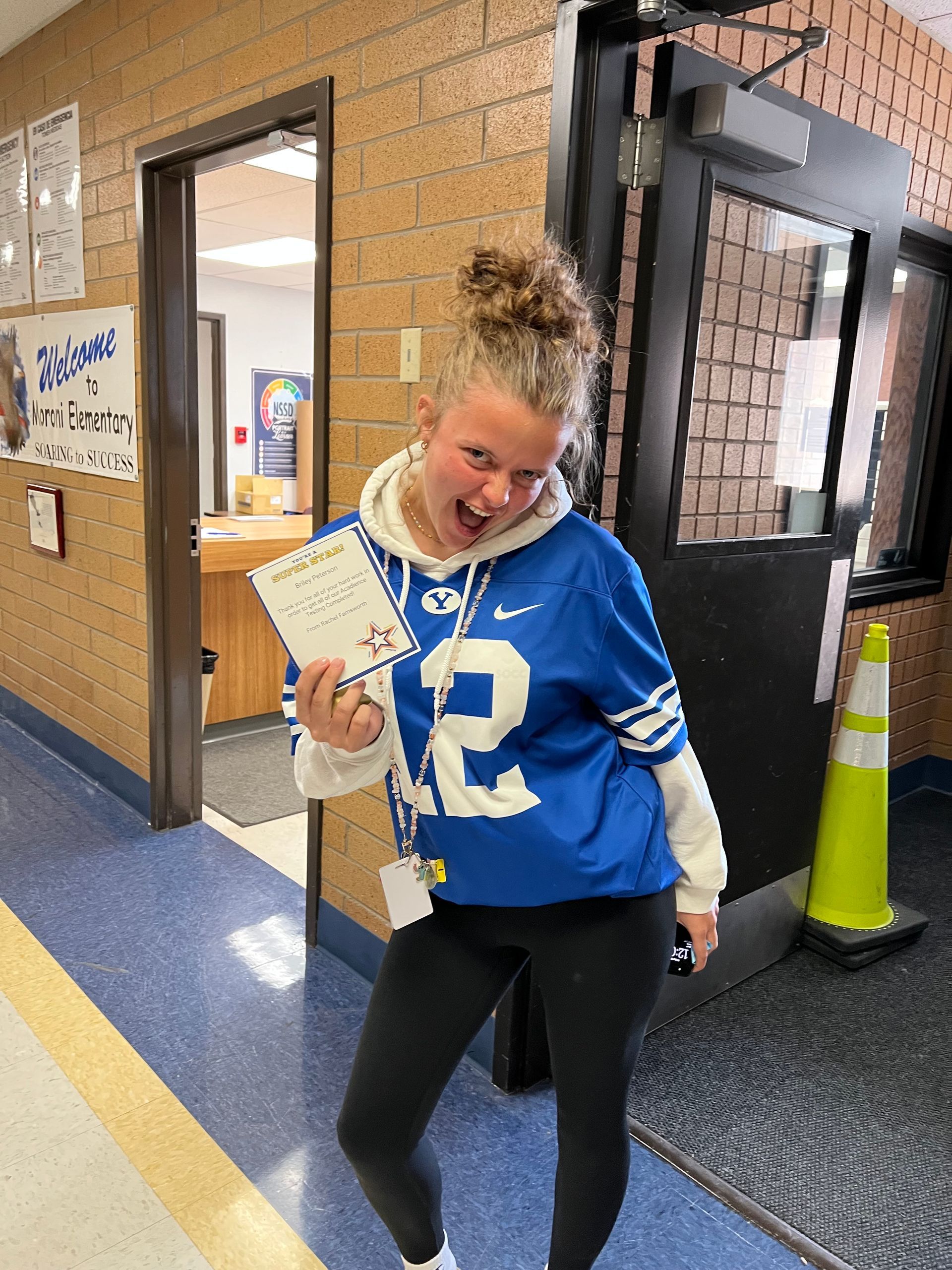 Girl in a BYU jersey holding a certificate winks in a school hallway.
