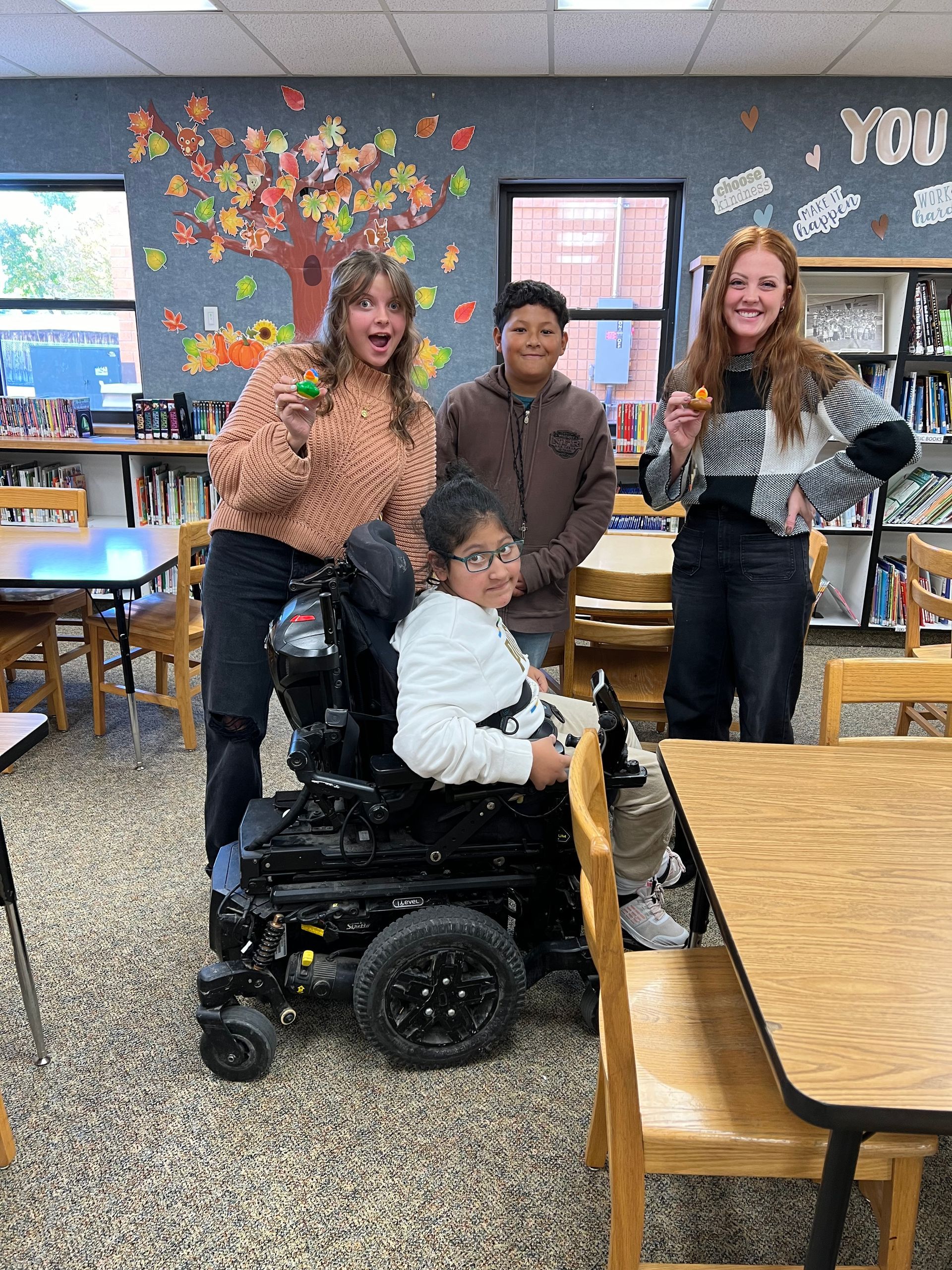 A group of people are standing around a boy in a wheelchair in a classroom.