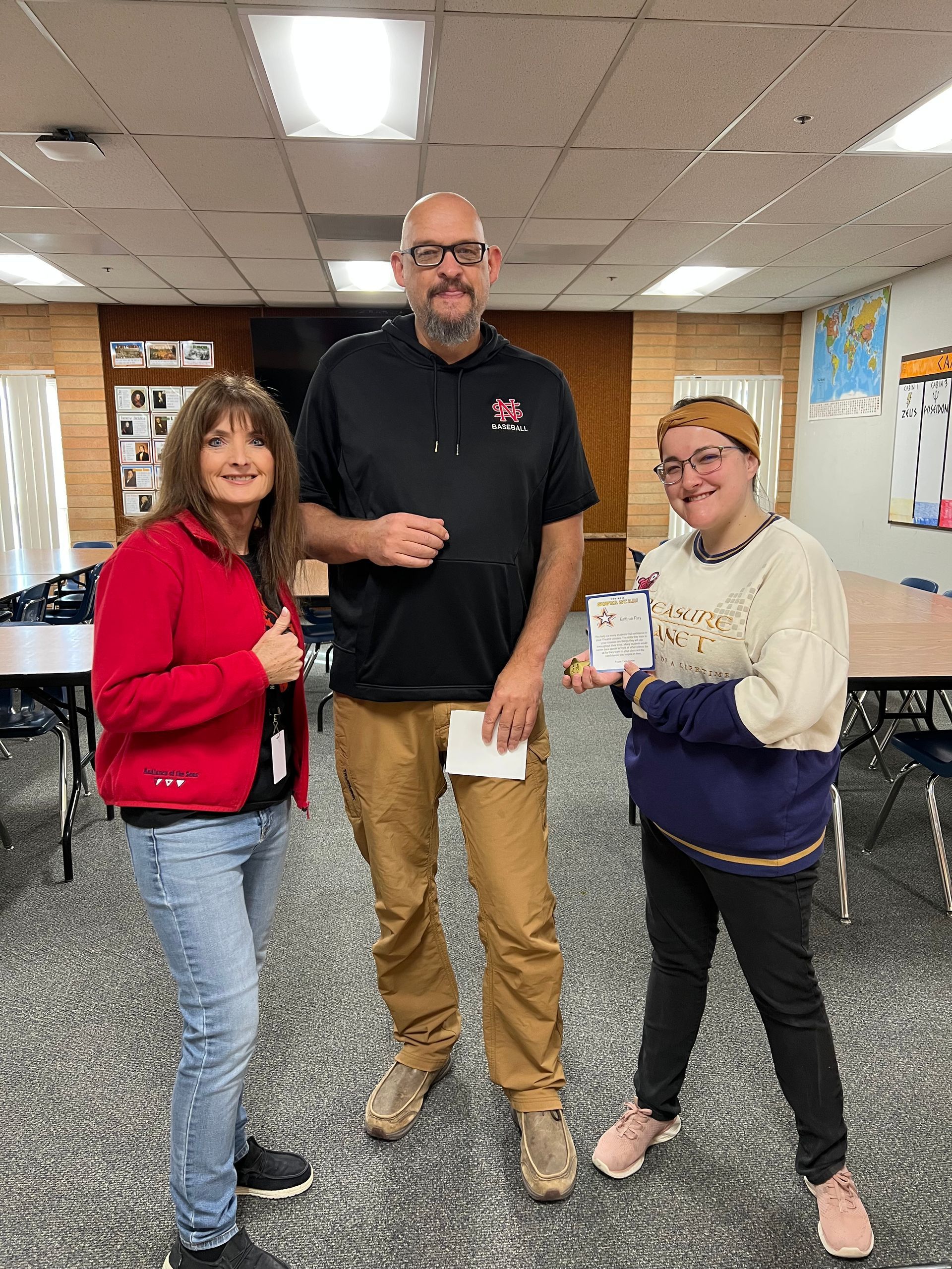 Three people in a classroom: Man in black sweatshirt holds a small box, flanked by woman in red and a woman in sweater.