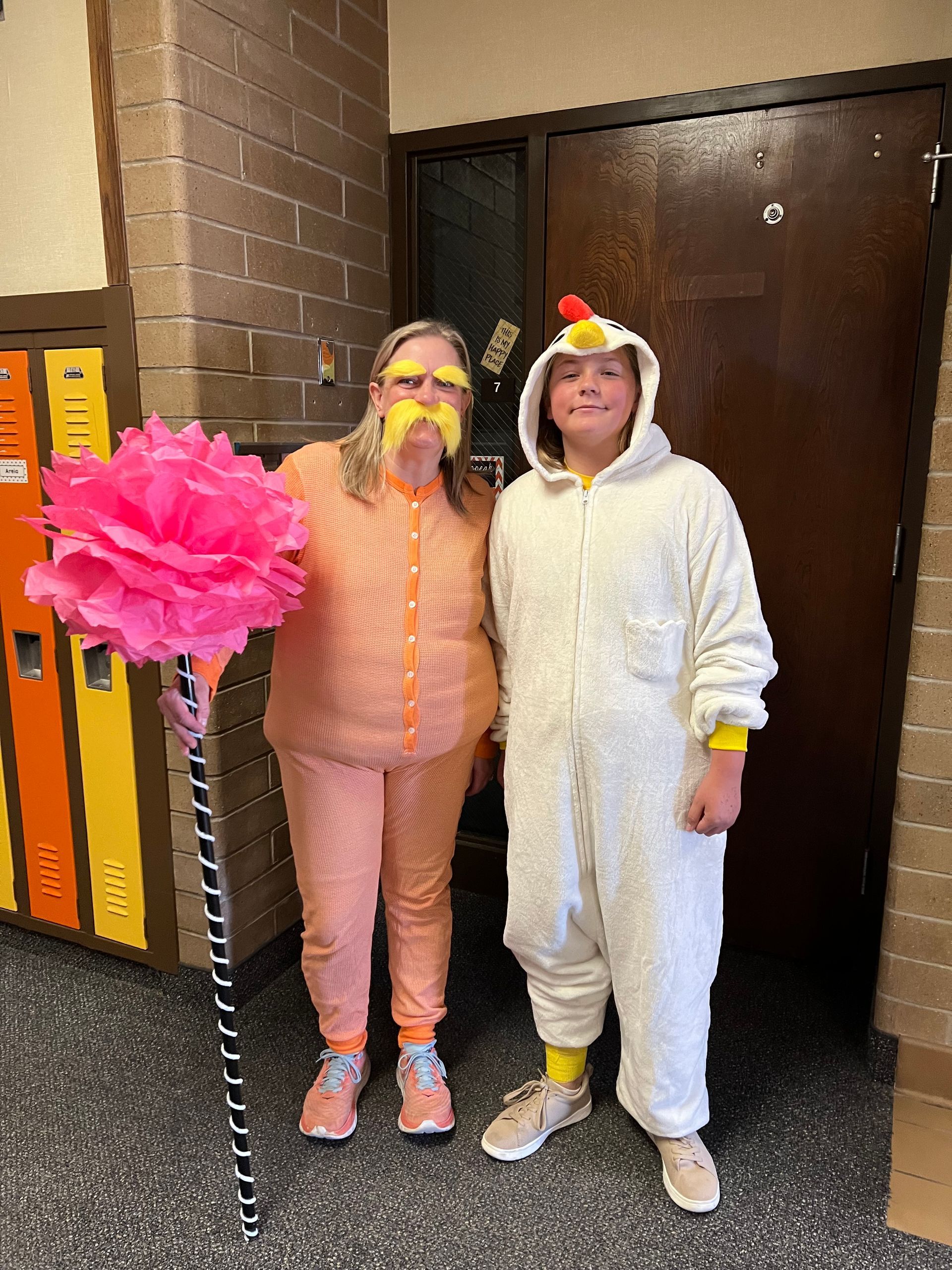 Person in orange Lorax costume with a pink Truffula tree and person in a chicken costume in a hallway.