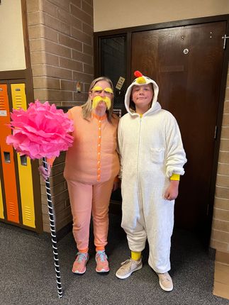 Woman in orange suit and child in chicken costume pose by lockers; woman holds pink flower.