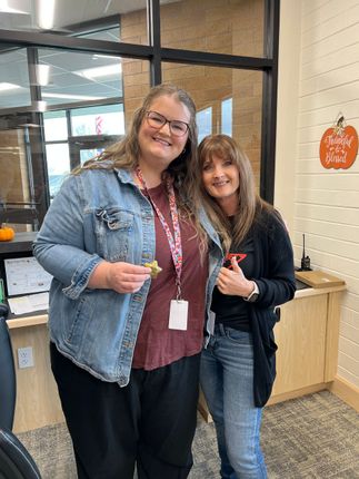 Two women smile, one in denim jacket, one in black sweater, inside office with window.