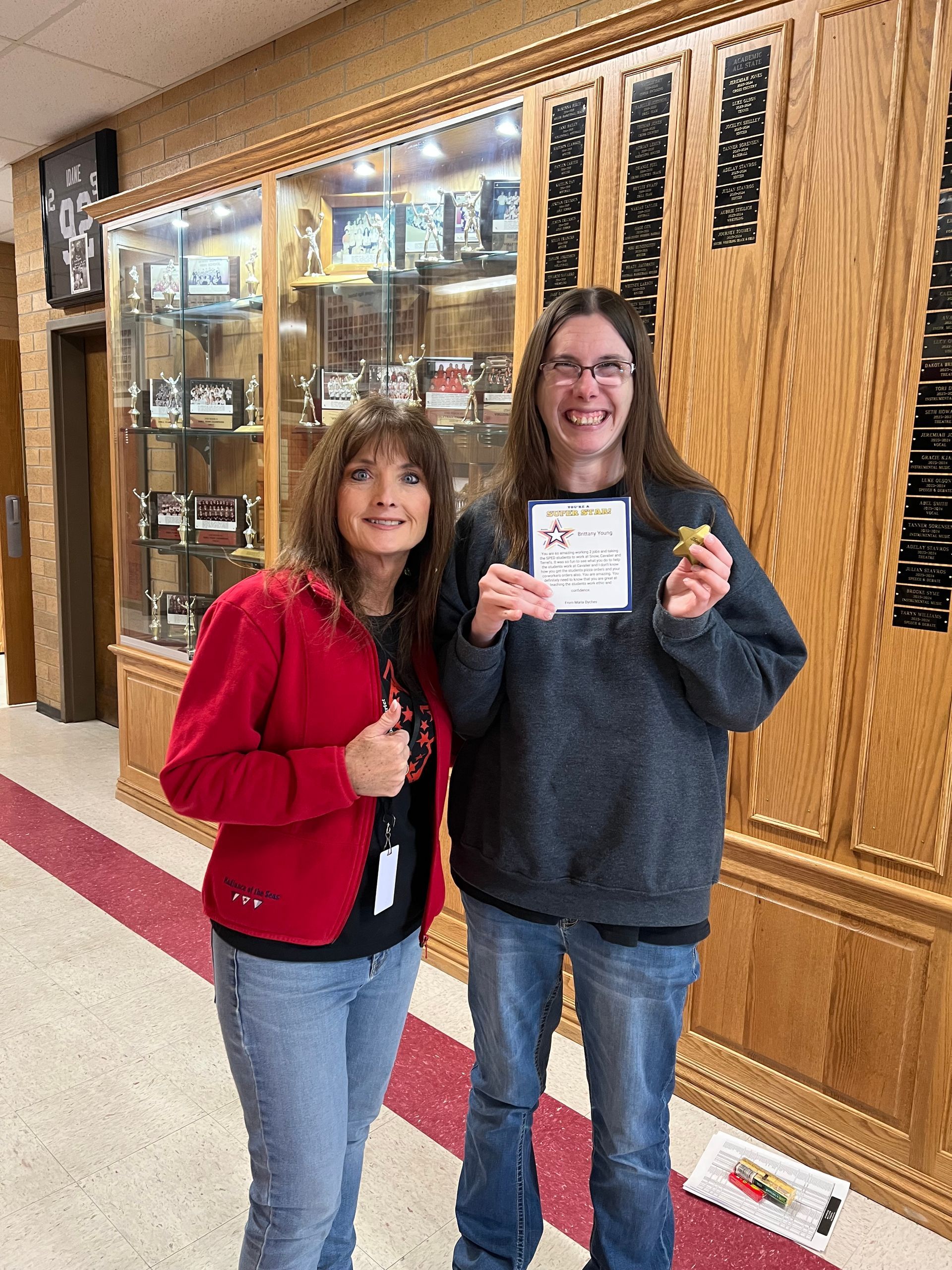 Woman in red jacket and person in gray sweater hold paper, smile. Display case in background.