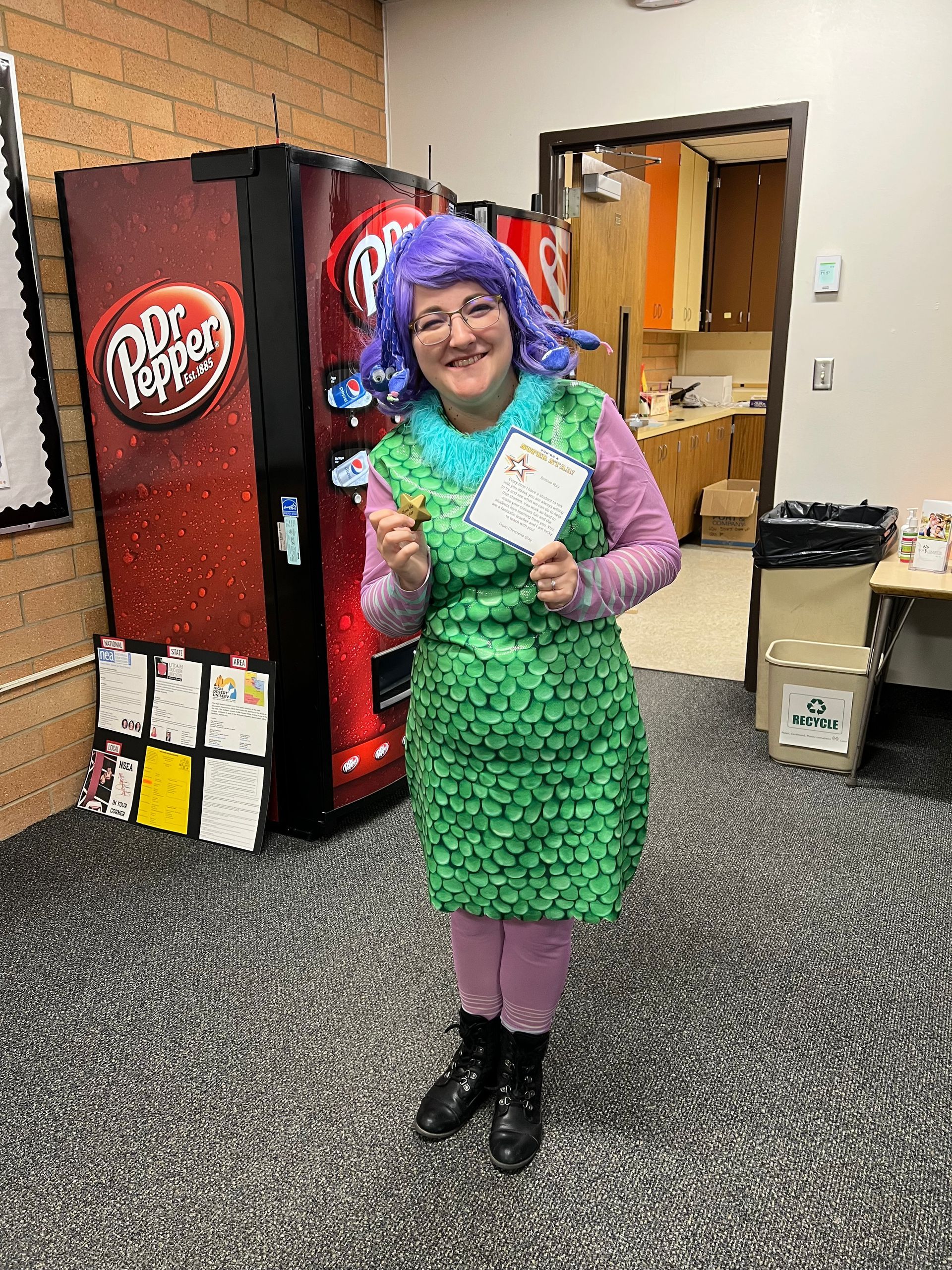 Person in a green scale costume and purple wig holding a card near a Dr. Pepper vending machine.