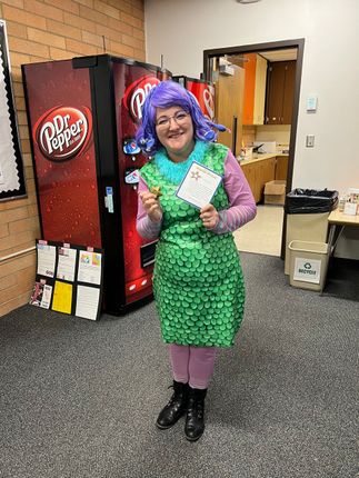 Person in a green scale costume and purple wig stands by a Dr. Pepper vending machine, holding a card.