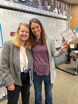 Two women smiling, posing in a classroom. One holds a small dry-erase board, the other has a name tag.