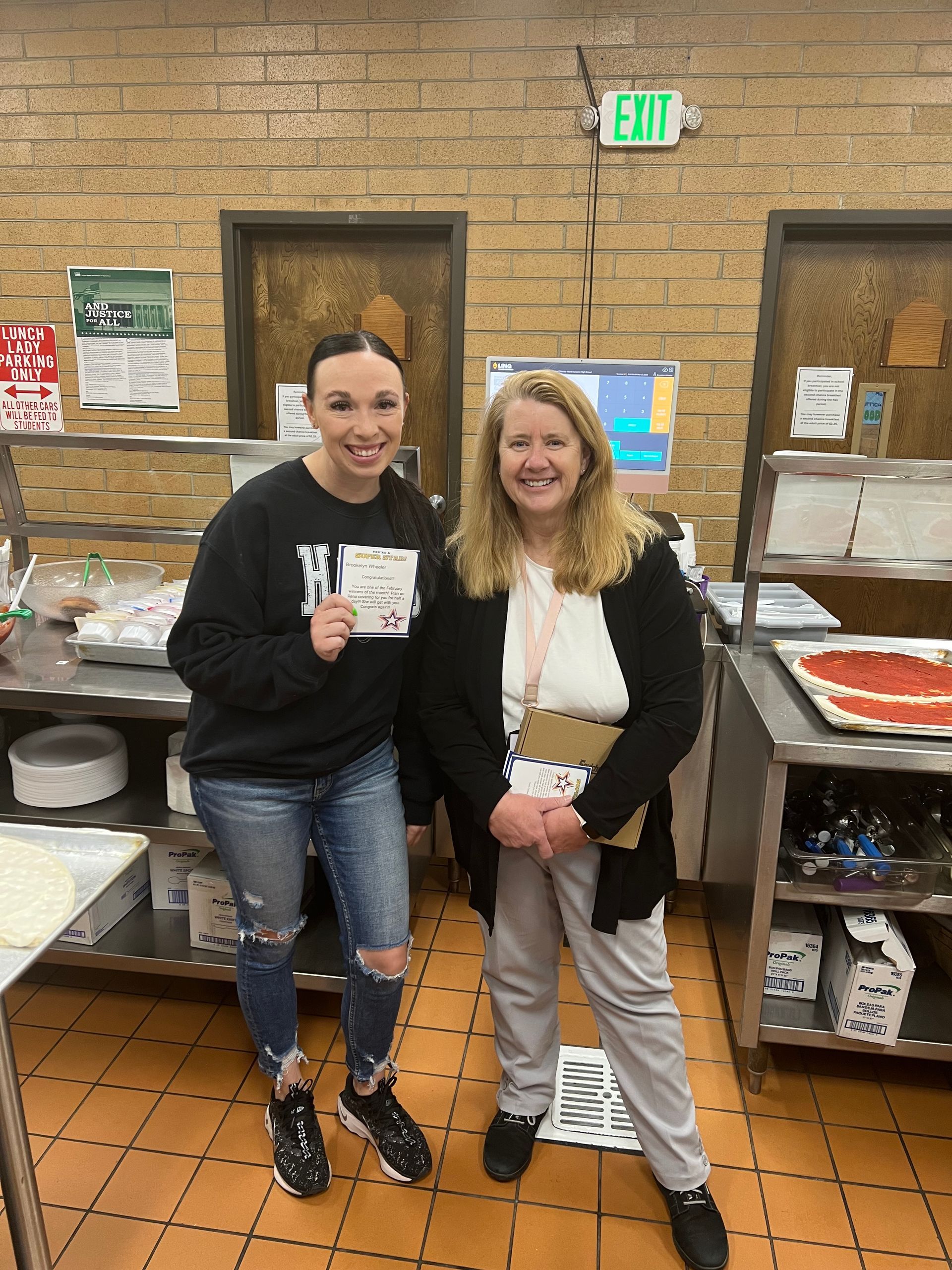 Two people stand together in a kitchen area, one holding a card, both smiling, with shelves and an exit sign in the background.