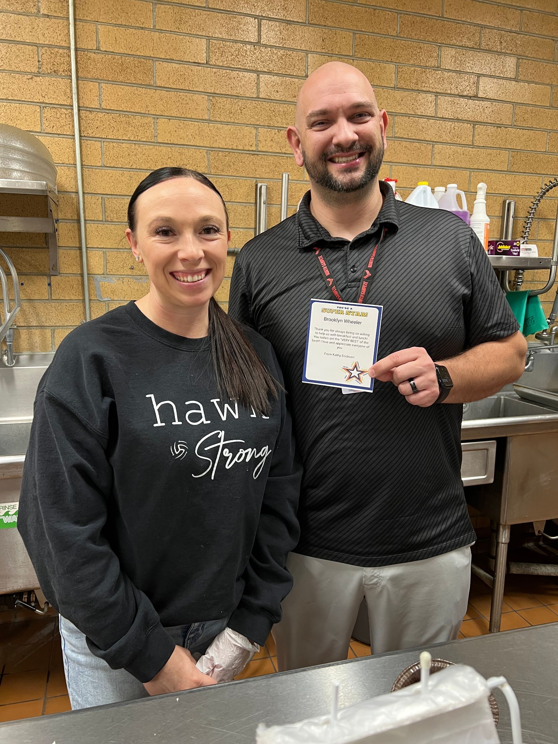 Two people smiling, posing in a commercial kitchen. Man holds a sign. Woman wears a 