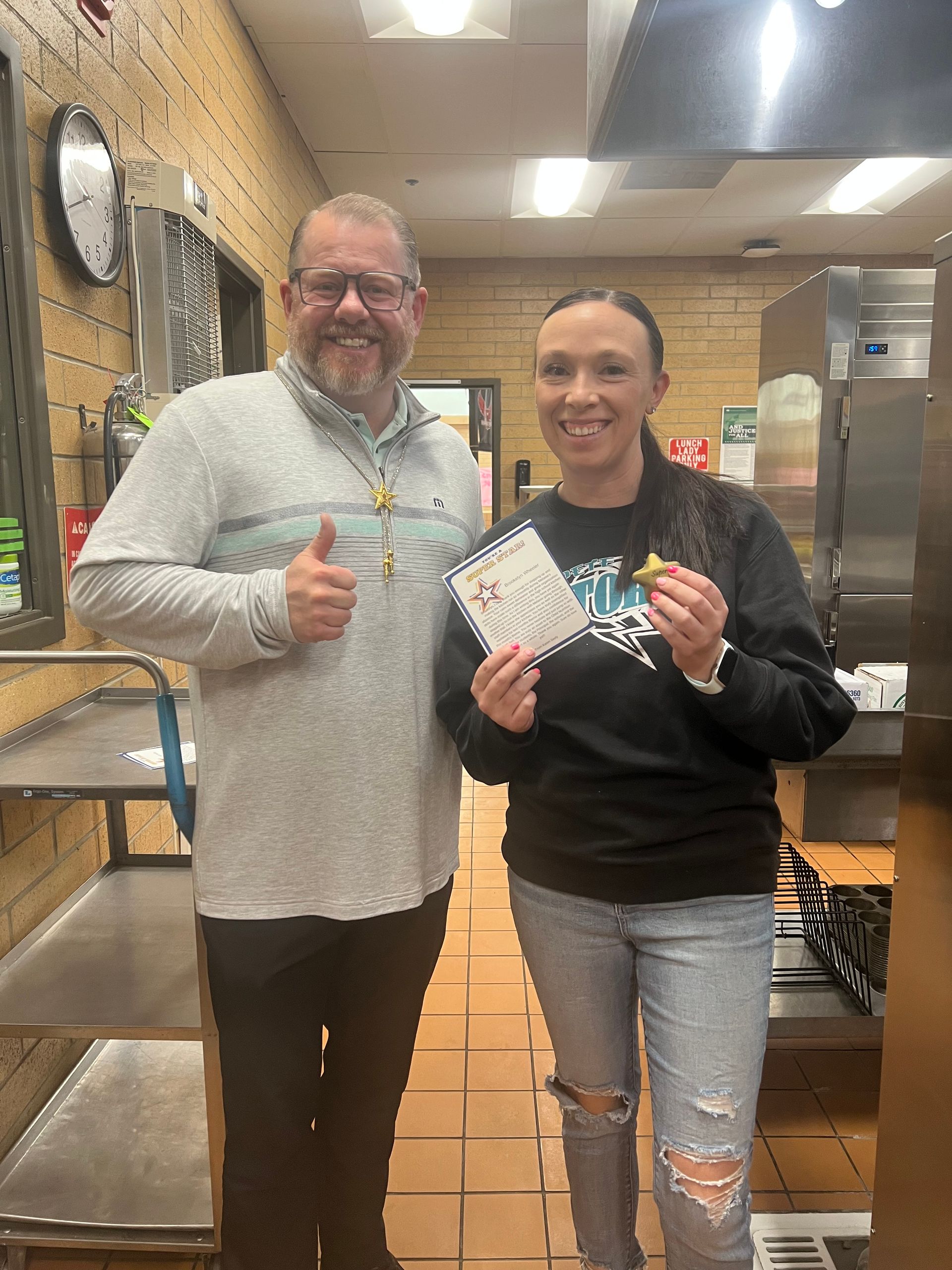 Man gives thumbs up, woman holds certificate and food item in a kitchen.