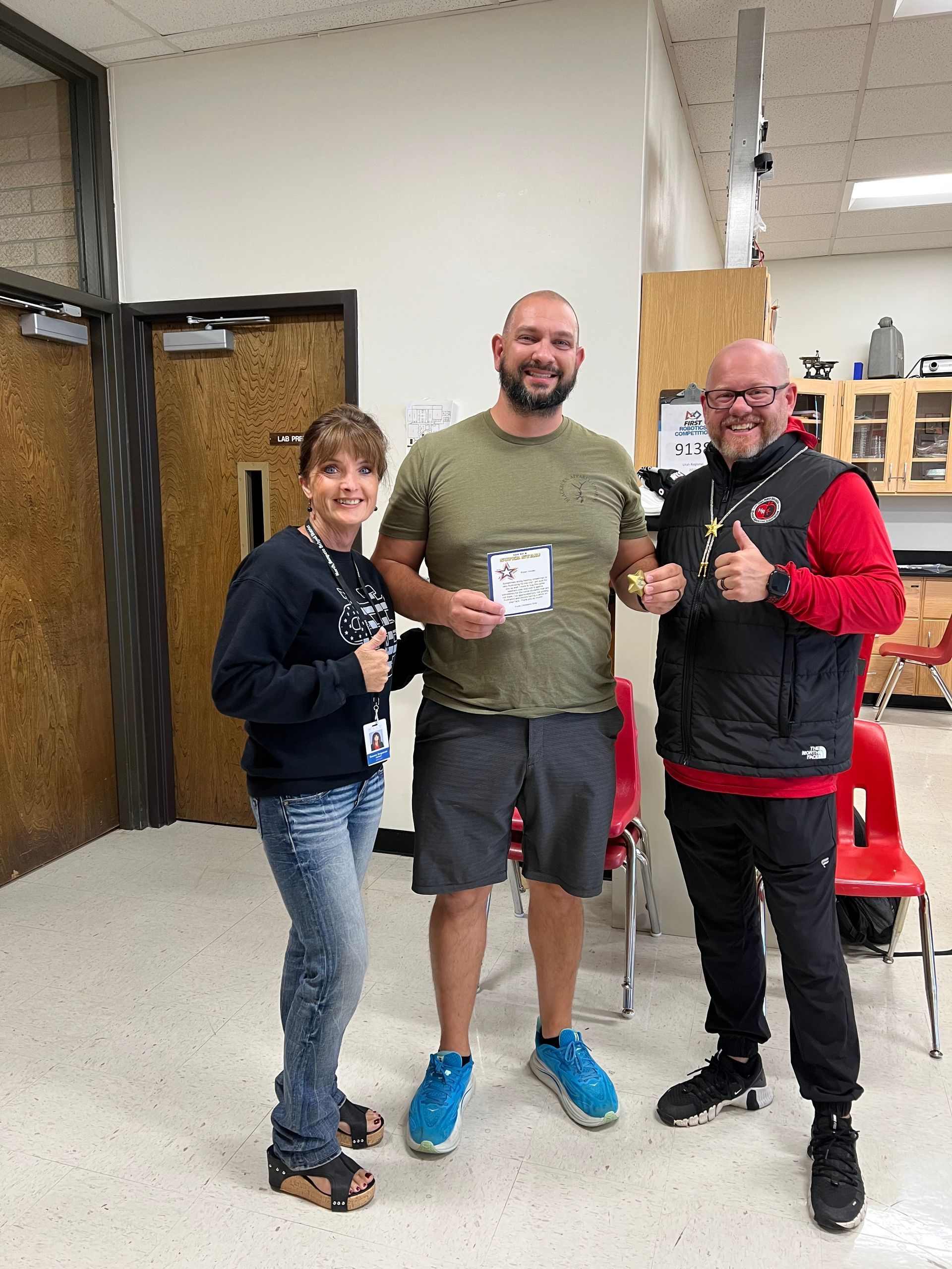 Three people pose with thumbs up in a classroom. The man in the center holds a certificate.