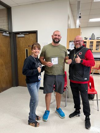 Three people give thumbs up. One holds a certificate. Indoors with red chairs, two doors, and some shelving.