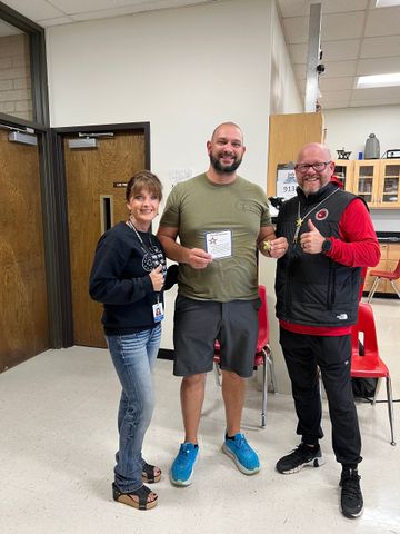 Three people give thumbs up. One holds a certificate. Indoors with red chairs, two doors, and some shelving.