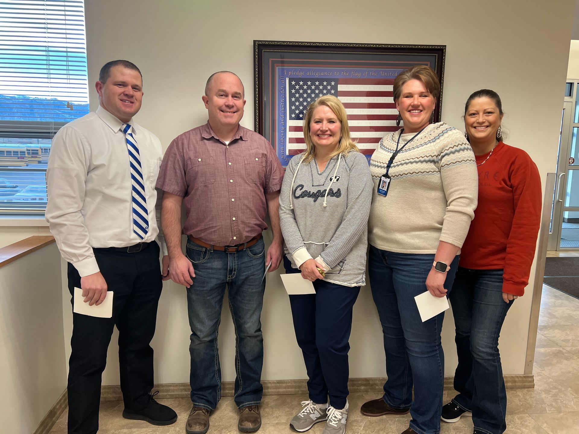 Five people stand indoors near a framed American flag. They hold papers, smiling, in a brightly lit hallway.