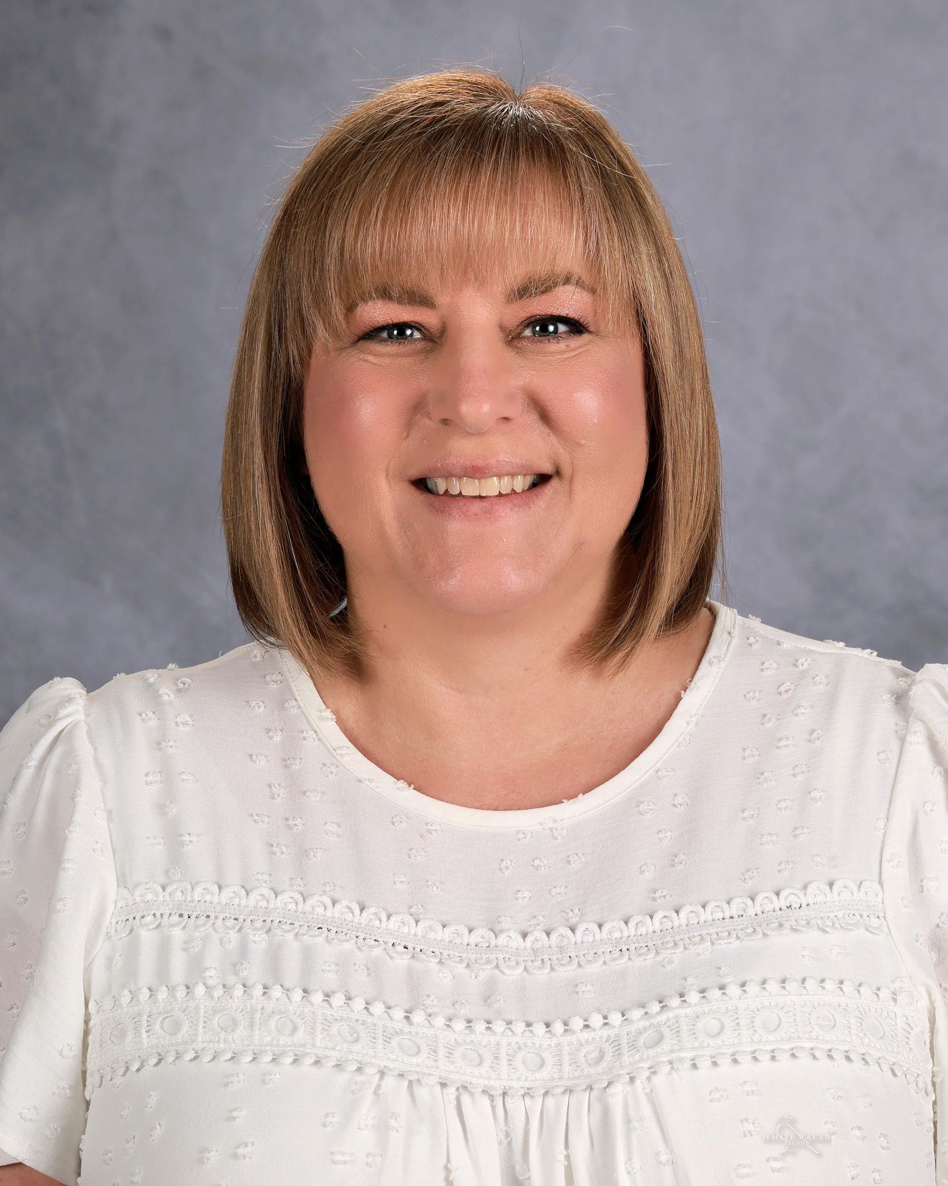 Woman with blonde hair and bangs smiles, wearing a white blouse, against a gray backdrop.