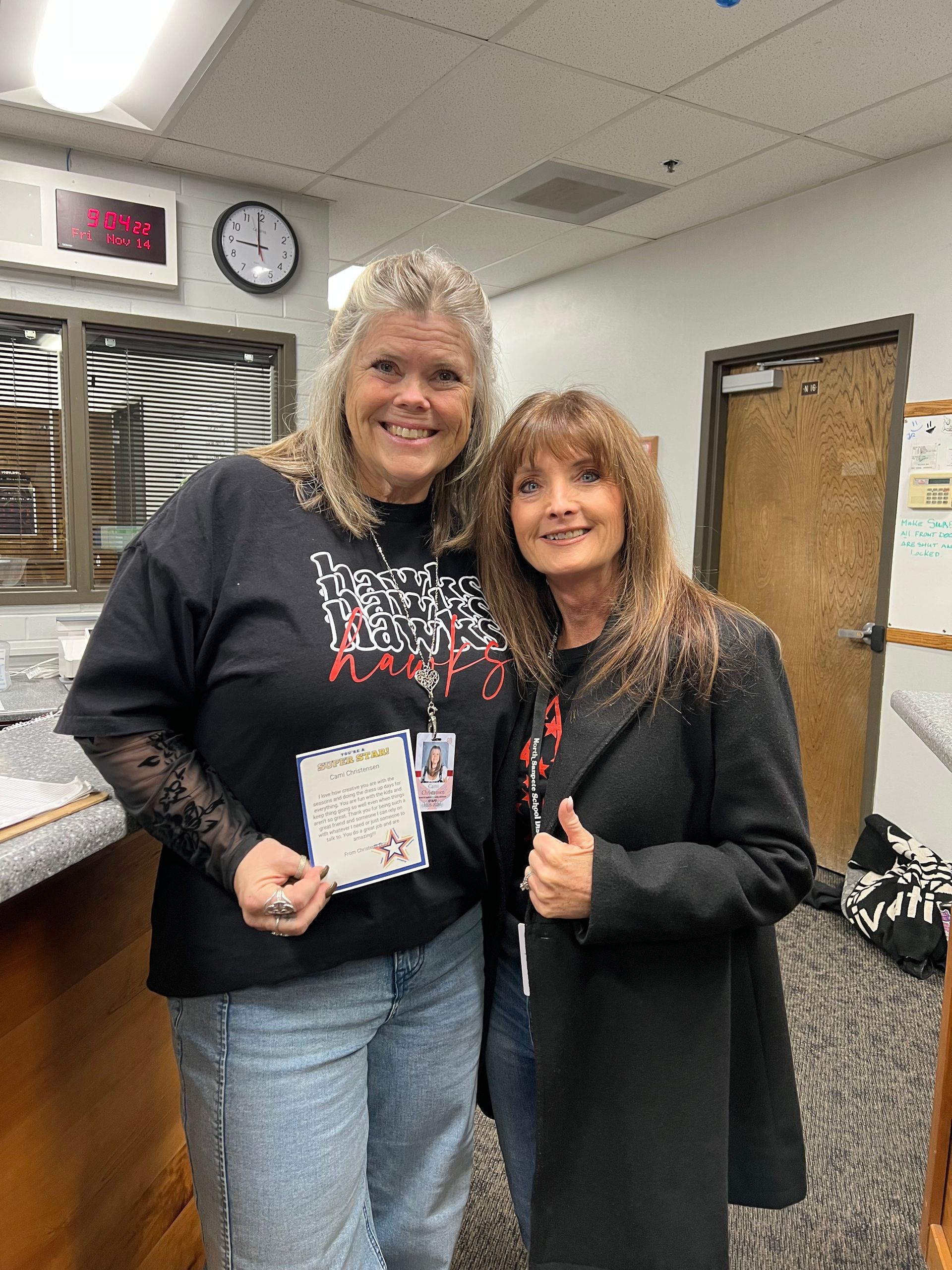 Two women smiling in an office. One holds a paper, wearing a black shirt and jeans. The other wears a black coat and jeans.