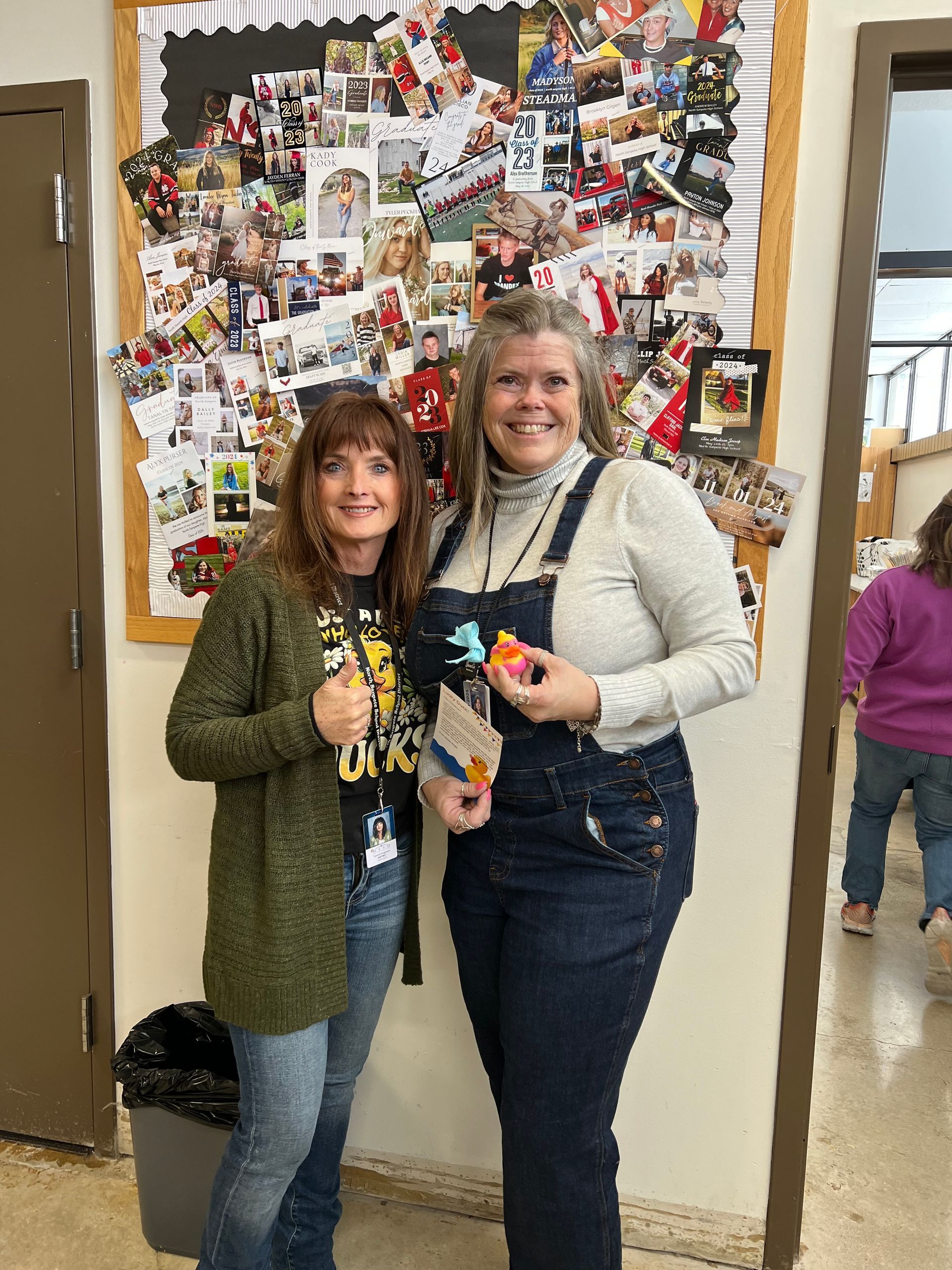 Two women are standing next to each other in front of a bulletin board.