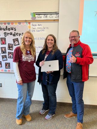 Three people smiling in a classroom, holding a laptop, with posters and a whiteboard behind them.