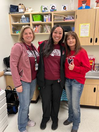 Three women standing together in a classroom, smiling and wearing maroon and red tops.