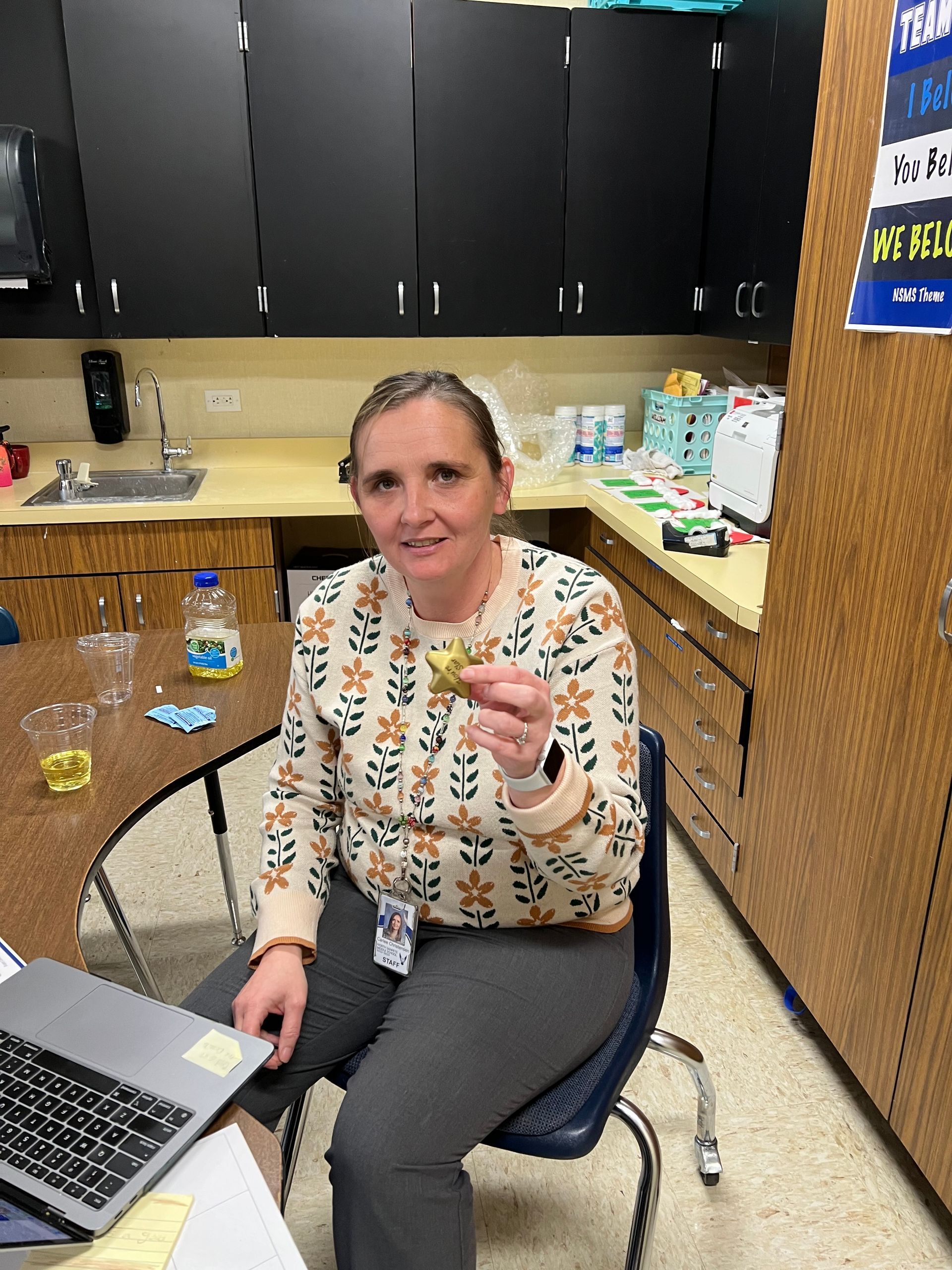 Woman holding a small item, seated at a desk in a classroom setting.