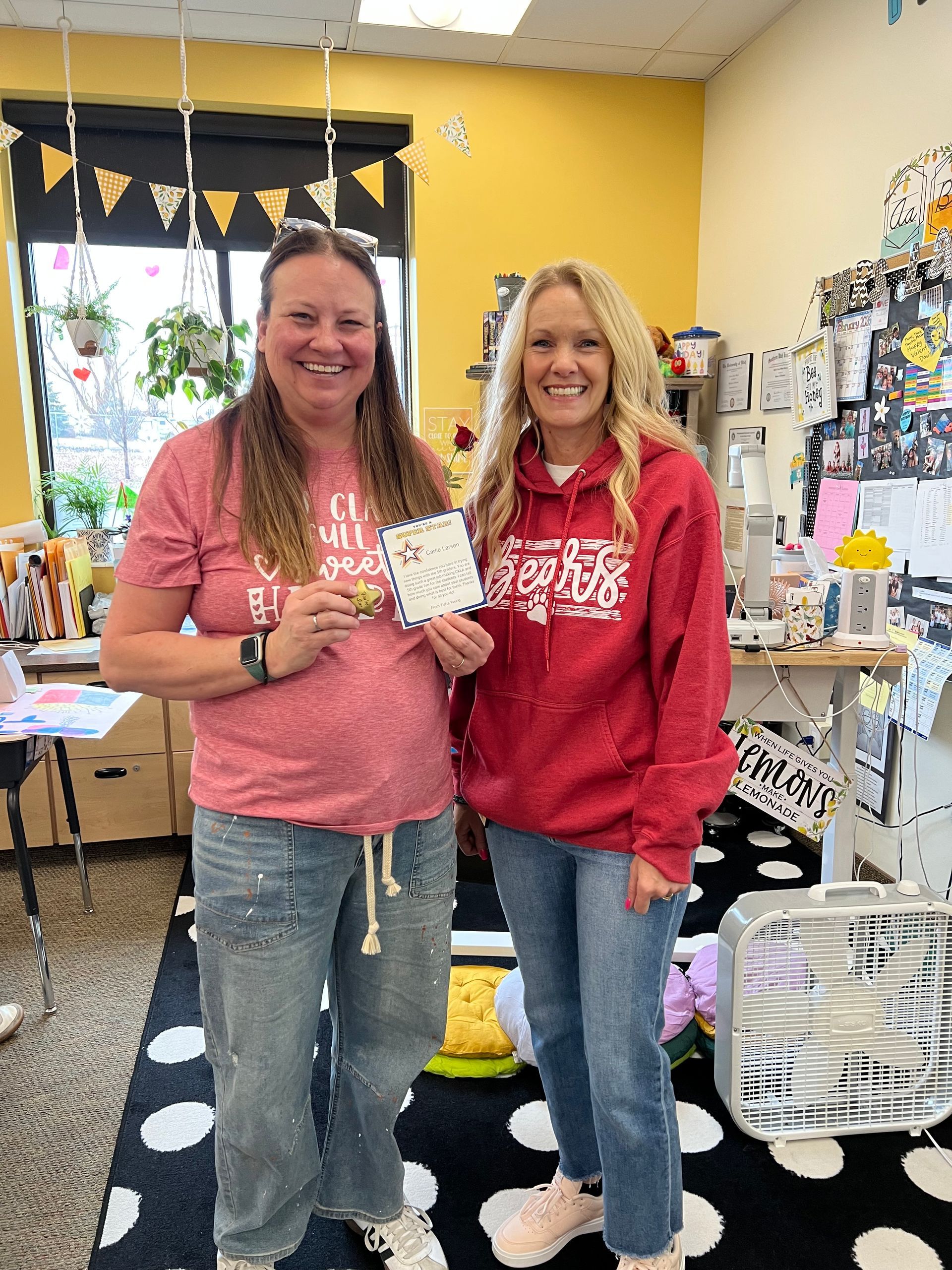 Two women in a classroom hold a craft. One wears red shirt and jeans, the other red hoodie and jeans.