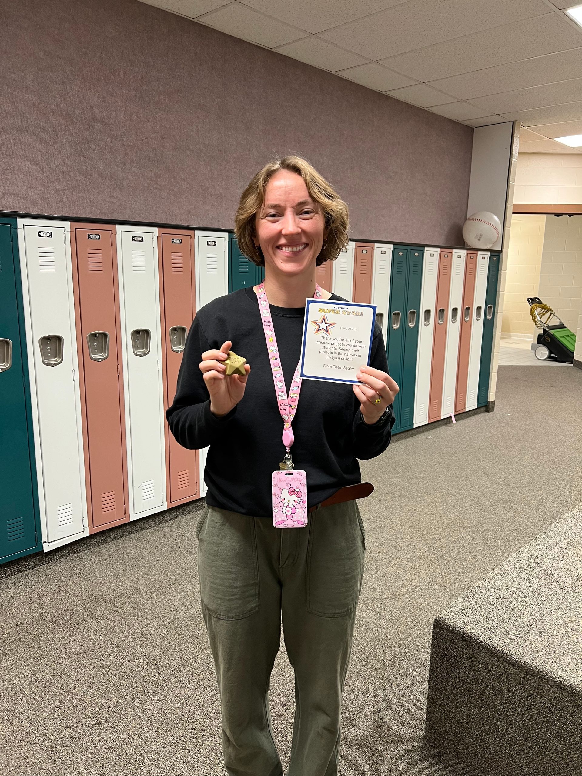 Woman in hallway holds a card and small item, smiles at the camera, wearing black shirt and green pants.