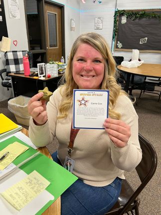 A smiling teacher holds a star-shaped object and a personalized award card at her desk in a classroom setting.