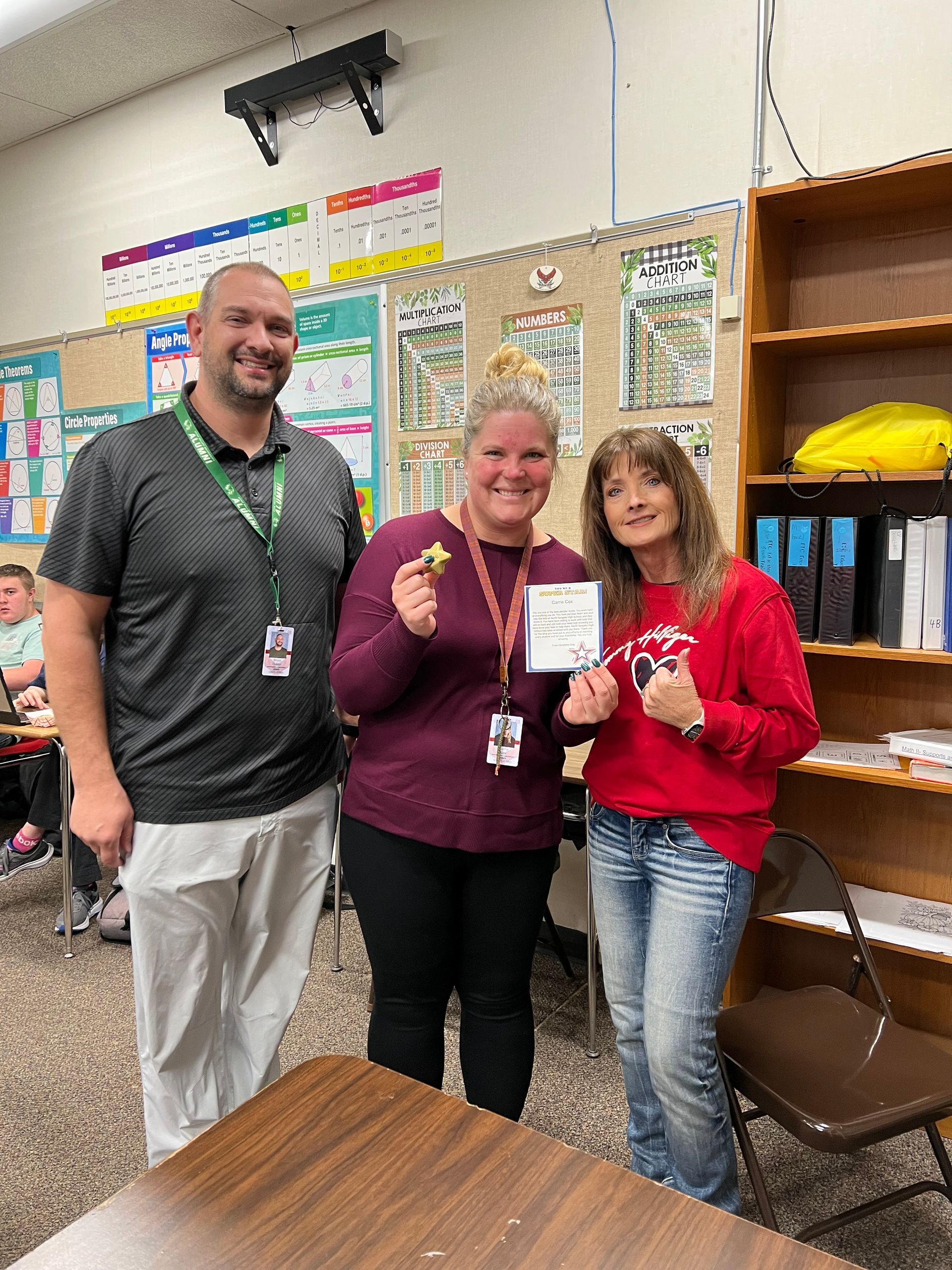 Three people smiling in a classroom, holding up a small card and badge, with posters and shelves behind them.