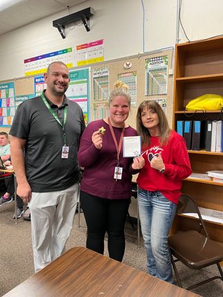 Three people smiling in a classroom, holding a small award card.