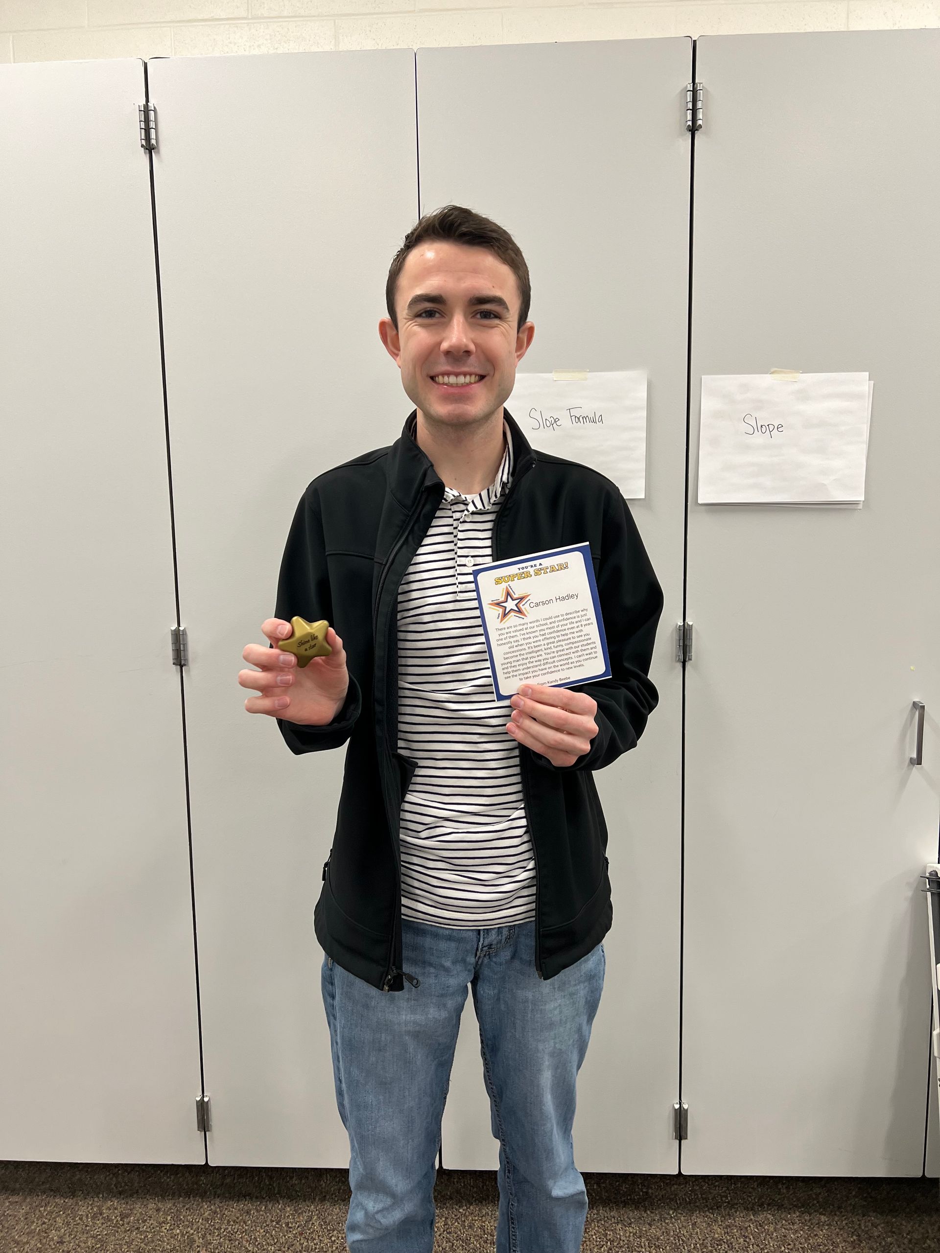 Man in black jacket and striped shirt holds a small item and a card, standing in front of a white wall.