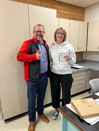Two people smiling and holding items in an office, with cabinets and a desk behind them.