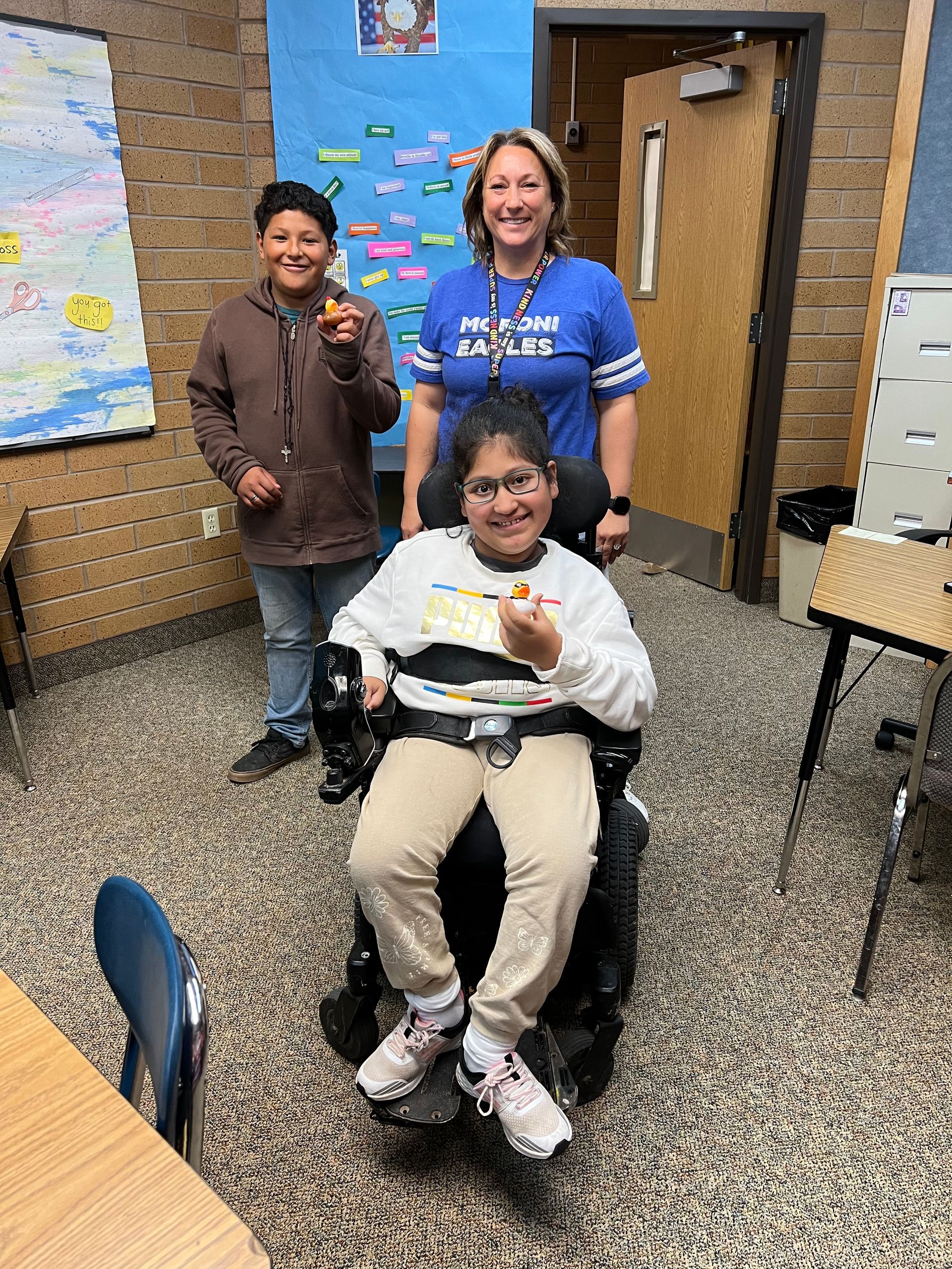 A woman is standing next to a girl in a wheelchair in a classroom.