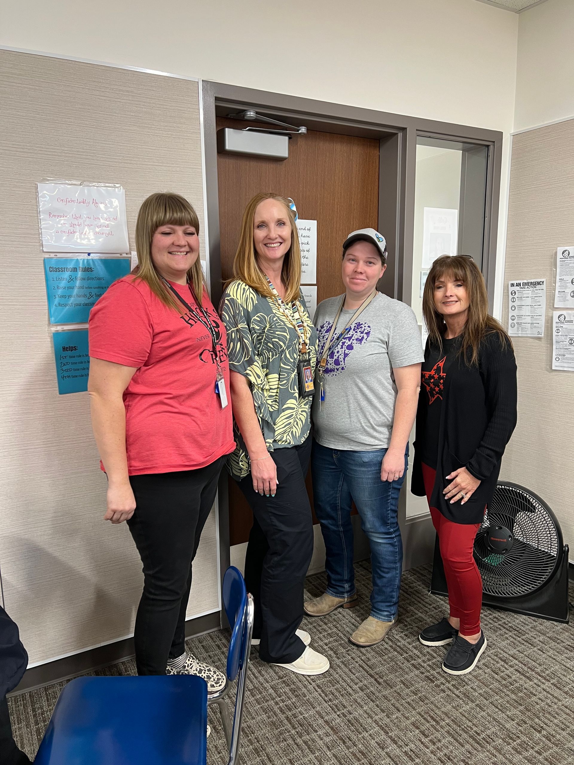 Four people stand in a doorway.  A woman in a red shirt, a woman in a floral shirt, a woman in a grey shirt, and a woman in a black shirt.