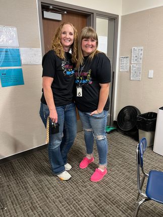 Two women smiling and posing indoors, wearing black shirts, jeans, and bright shoes.