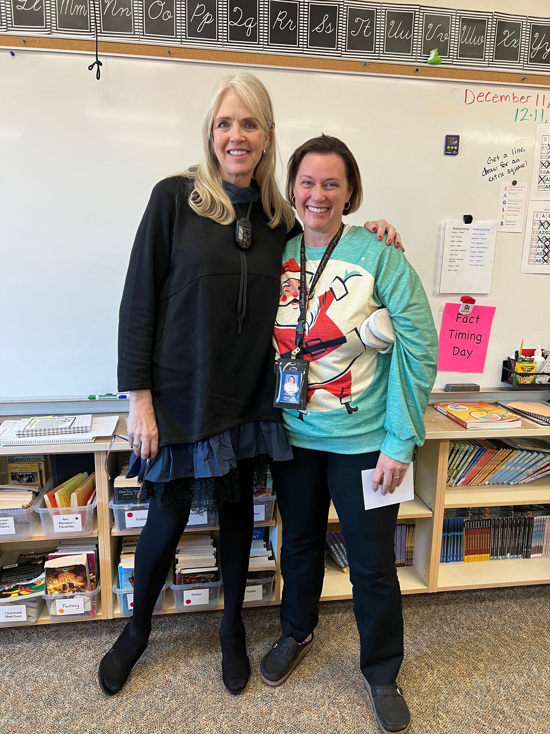 Two women stand, smiling, in a classroom. One wears a black sweater, the other a Santa sweater. Whiteboard in background.