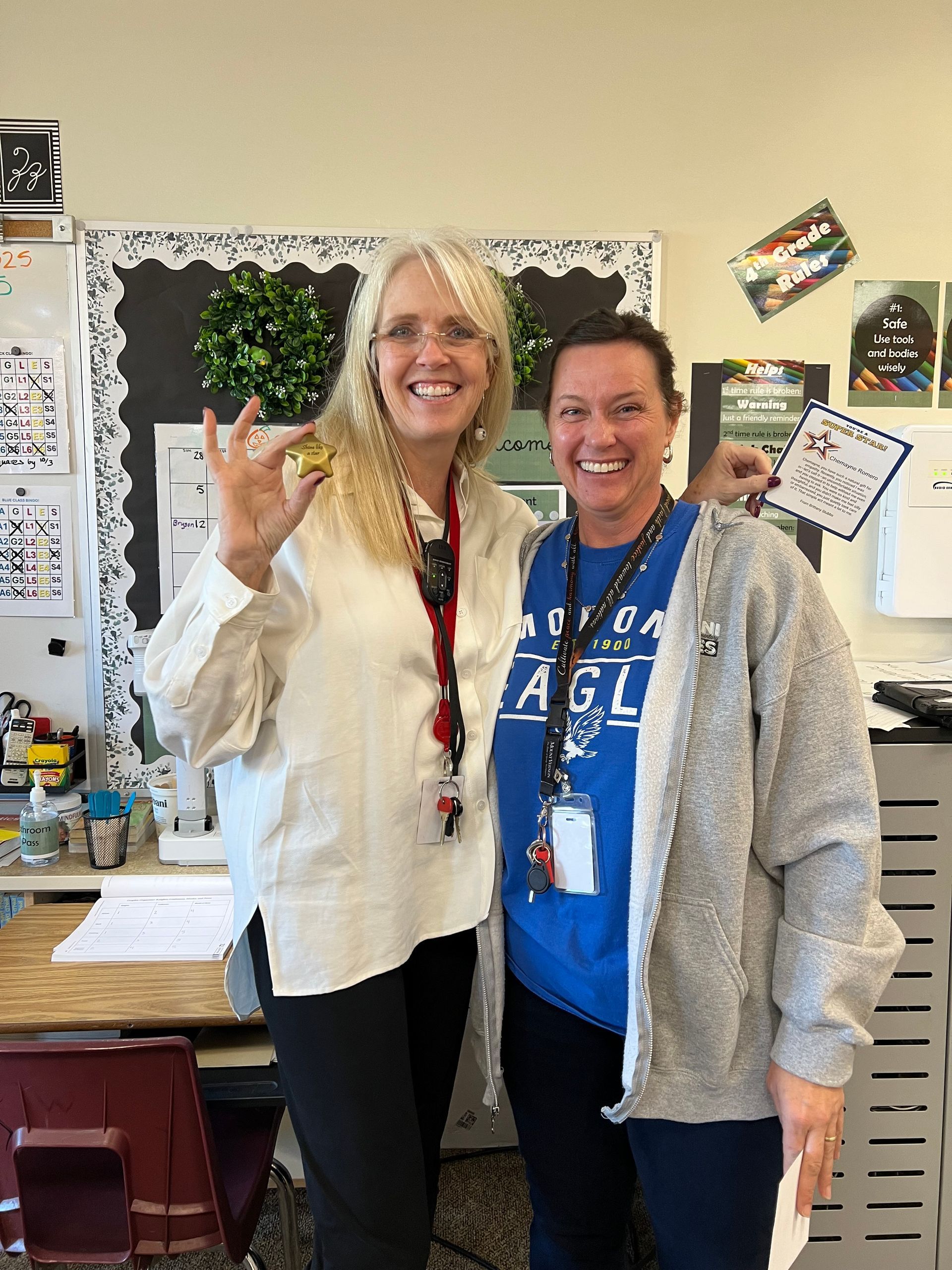 Two women smiling in a classroom, one holding up a gold bell.