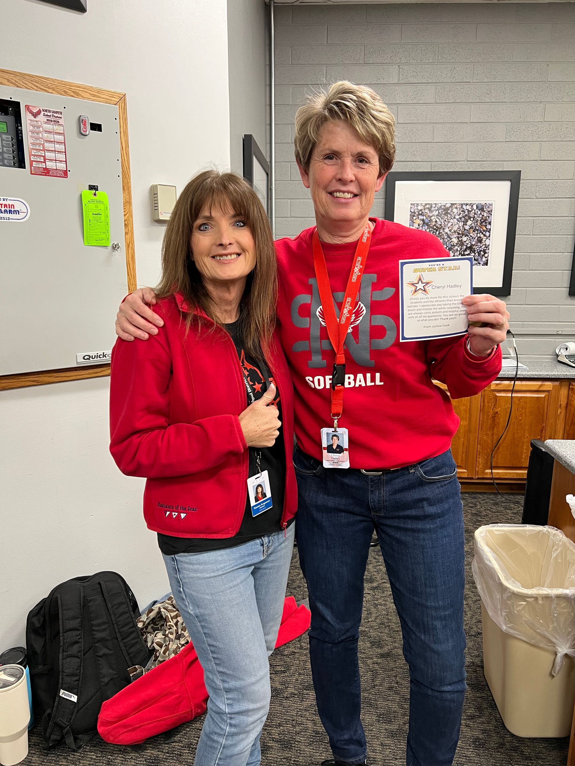 Two women pose, one holding a certificate, both wearing red sweatshirts and jeans.