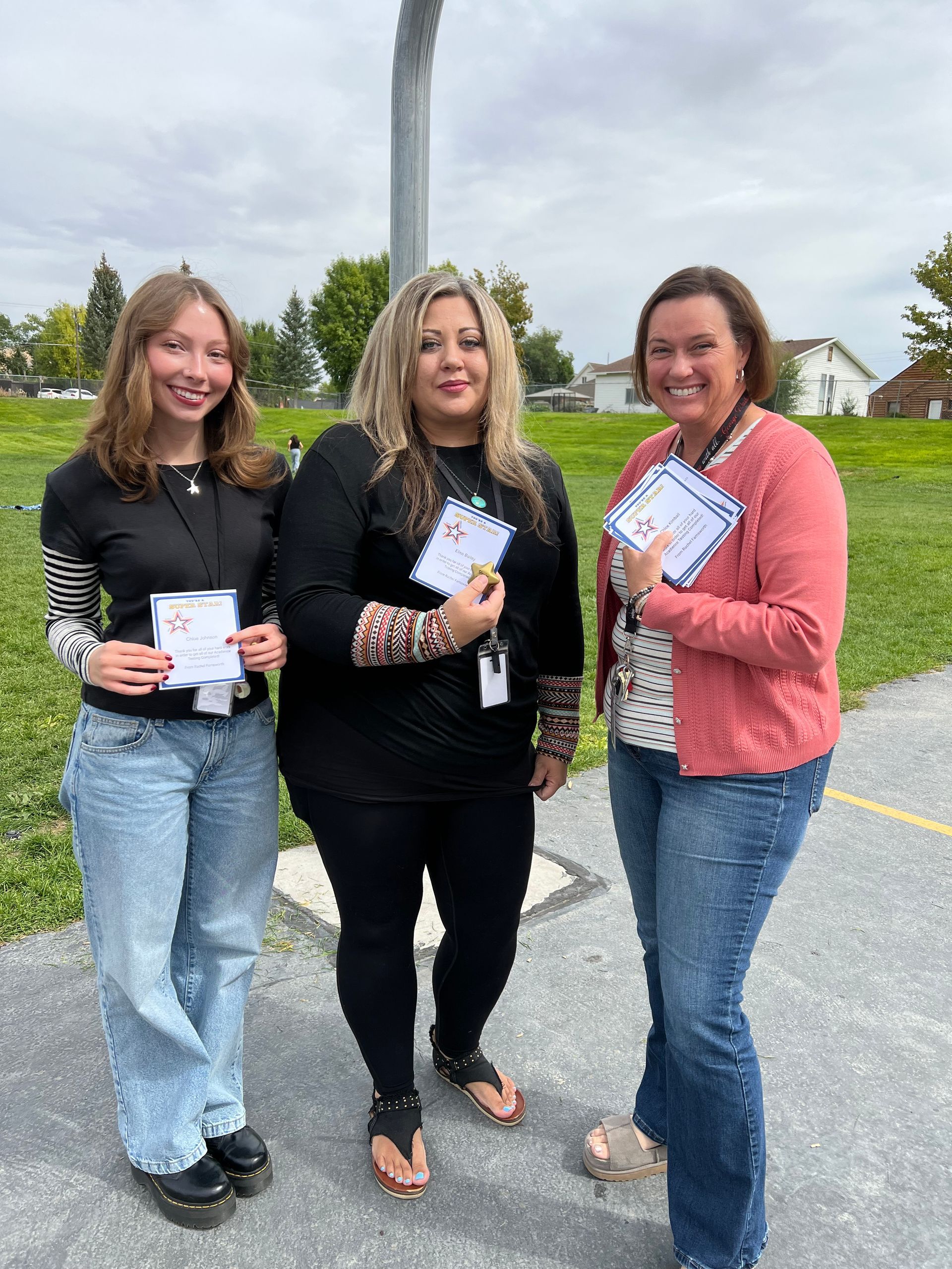 Three women holding papers outdoors. Smiling. Left: blonde, jeans, striped sleeves. Middle: dark hair, black clothing. Right: brown hair, pink sweater.